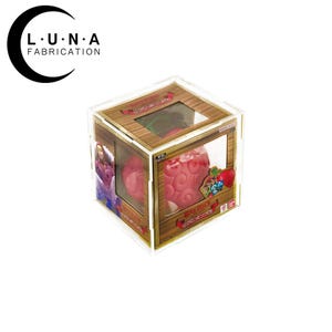 May include: A pink, textured fruit-shaped object inside a clear acrylic display box. The box has a wooden-style frame and a window showing the object. The box has a One Piece logo and other Japanese text.
