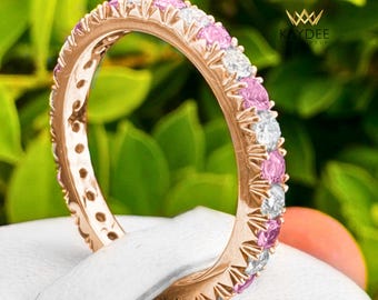 Könnte beinhalten: Ewigkeitring aus Roségold mit abwechselnd rosa und weißen Edelsteinen. Der Ring ist vor einem Hintergrund aus grünem Laub platziert, mit dem Kaydee-Logo oben rechts. Das Design des Rings zeigt eine durchgehende Reihe von Edelsteinen.