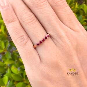 Könnte beinhalten: Ein roségoldener Ring mit sieben roten Edelsteinen in einer Reihe. Der Ring wird auf einer Hand gezeigt, mit einem verschwommenen grünen Hintergrund. Der Ring ist ein zarter Schmuck.