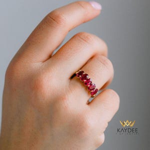 May include: A gold ring with five oval-cut, vivid red gemstones. The ring is set in a gold band and is shown on a hand. The jewelry is from Kaydee Fine Jewelry.
