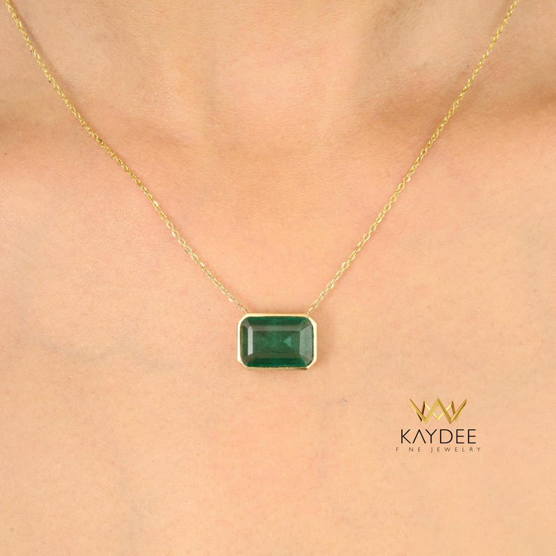 Op de afbeelding: Een gouden ketting met een rechthoekige, smaragdgroene edelsteen hanger. De hanger is gezet in een gouden bezel en hangt aan een fijne gouden ketting. De ketting wordt getoond tegen een neutrale achtergrond.