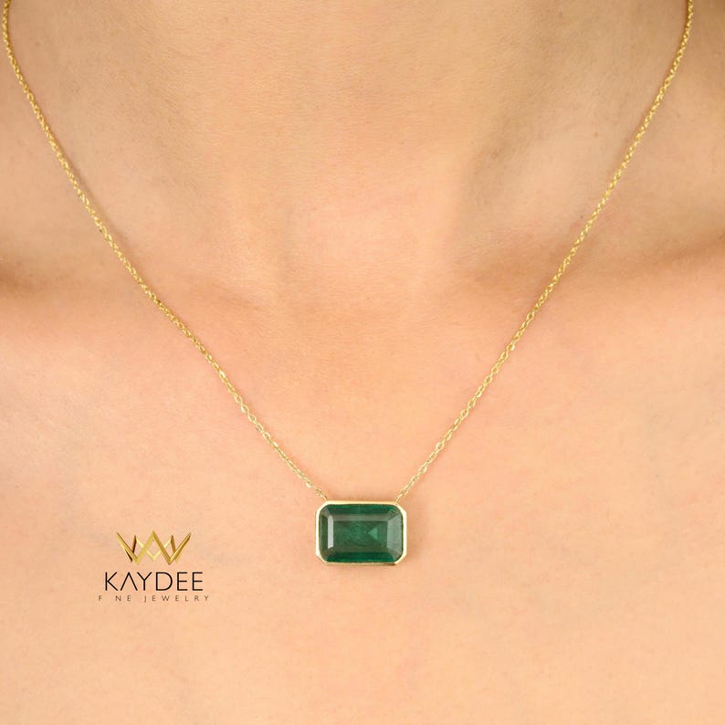 Op de afbeelding: Een gouden ketting met een rechthoekige, smaragdgroene edelsteen hanger. De hanger is gezet in een gouden bezel en hangt aan een fijne gouden ketting. De ketting wordt om de nek van een persoon gedragen. De merknaam "KAYDEE FINE JEWELRY" is zichtbaar.