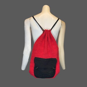 May include: Red crocheted drawstring backpack with a black crocheted pocket and two black buttons.