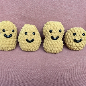 Könnte beinhalten: Vier handgefertigte, plüschige, gelb-braune Häkel-Lebensmittel, die Hähnchen-Nuggets ähneln. Jedes hat schwarze Knopfaugen und ein schwarzes, gebogenes Lächeln. Die Artikel sind auf einer rosa Oberfläche angeordnet.