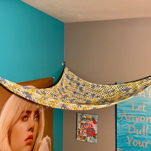 May include: A yellow, blue, and white crocheted hammock hanging from the ceiling in a room with teal walls. The hammock is suspended from two hooks on the wall. There are three framed pictures on the wall behind the hammock.