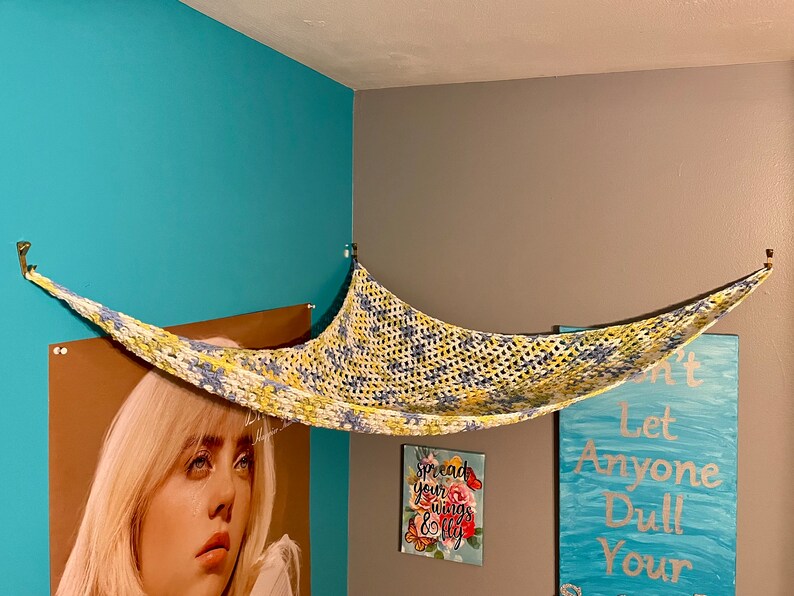 May include: A yellow, blue, and white crocheted hammock hanging from the ceiling in a room with teal walls. The hammock is suspended from two hooks on the wall. There are three framed pictures on the wall behind the hammock.