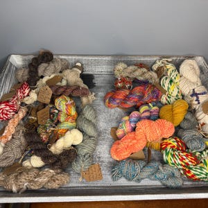 May include: A wooden tray filled with various colorful yarn skeins. The yarn colors include red, orange, yellow, green, blue, and brown. Some skeins have small tags attached. The tray has a distressed, white-washed finish.
