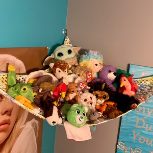 May include: A colorful crocheted hammock filled with a variety of plush toys, including a green frog, a brown dog, a purple bear, and a pink flamingo. The hammock is hanging from the ceiling and is decorated with a yellow and blue pattern.