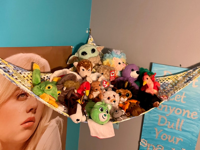 May include: A colorful crocheted hammock filled with a variety of plush toys, including a green frog, a brown dog, a purple bear, and a pink flamingo. The hammock is hanging from the ceiling and is decorated with a yellow and blue pattern.
