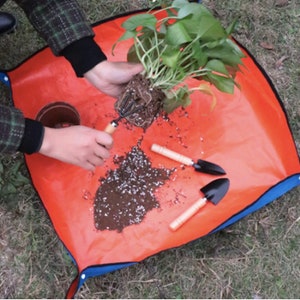 Op de afbeelding: Een vierkante oranje waterdichte tuinmat met blauwe drukknopen op de hoeken, met een groene potplant met blootliggende wortels, een kleine bruine plastic pot, een handhark met drie tanden en twee kleine zwarte metalen schepjes met houten handvatten.