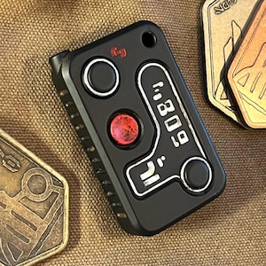 May include: Black rectangular device with two circular buttons and a red button. The device has the text "809" and other symbols in white. Several bronze-colored metal coins are in the background.