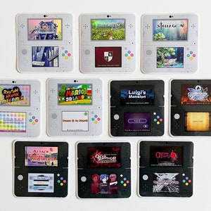 May include: Nine different Nintendo 3DS game cartridge stickers. The stickers are white and black with images of the game covers. The games are: Rhythm Heaven Megamix, Super Mario 3D Land, Luigi's Mansion, Fire Emblem: Awakening, Stella Glow,  Attack on Titan: Humanity in Chains, Devil Survivor 2: Record Breaker,  Code of Princess, and  Fire Emblem: Shadow Dragon.