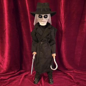 May include: A doll featuring a pale white face with large black eye sockets, wearing a black fedora, a long black trench coat with a belted waist, and black trousers. The doll has a silver-colored blade on the right hand and a silver hook on the left.