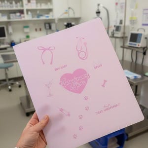May include: A light pink folder with various veterinary-themed graphics, including a bow, stethoscope, martini glass, syringe, bone, and paw prints. The text "Veterinary Girls" is inside a pink heart. Additional text includes "Dog Lady", "Spicy", and "Cat Whisperer".