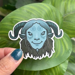 May include: A sticker featuring a blue-skinned creature with curved horns, a smiling face, and dark hair. The sticker has a white border and is held against a backdrop of green leaves.