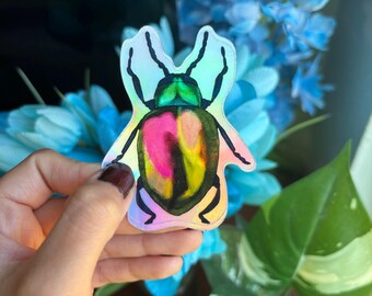 May include: A holographic sticker of a colorful beetle with green, pink, and yellow accents. The beetle is depicted with its wings spread out.