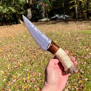 May include: A knife featuring a translucent, light blue, flaked stone blade with a sharp, pointed tip. The handle is crafted from a textured, cream-colored antler base with a dark brown, polished wooden bolster section near the blade.