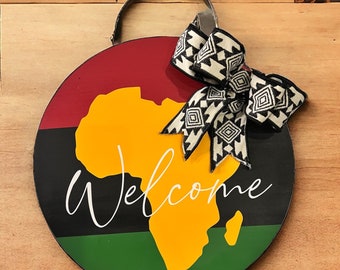 May include: A round wooden door hanger with a black, red, green, and yellow color scheme. The hanger features a silhouette of the continent of Africa and the word "Welcome". A black and white patterned bow is attached to the top of the hanger.