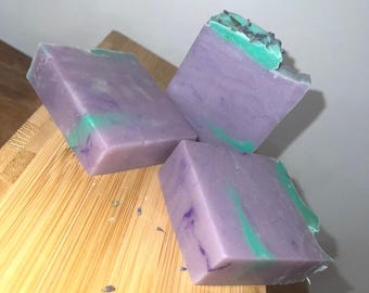 May include: Three rectangular bars of handmade soap. The soap is a lavender color with streaks of green and purple. The soap is sitting on a wooden cutting board.