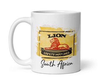 Könnte beinhalten: Weiße Keramiktasse mit einer Grafik im Vintage-Stil einer Löwen-Streichholzschachtel. Die Grafik zeigt eine gelbe Schachtel mit schwarzem Text, der "Lion Safety Matches" lautet, und eine rote Löwenillustration. Der Text "South Africa" ist unter der Grafik gedruckt.