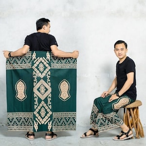 Puede incluir: Dos hombres vistiendo un sarong de patrón verde y crema. El sarong tiene un diseño geométrico con un motivo central de media luna y estrella. Un hombre está sosteniendo el sarong para mostrar el patrón, mientras que el otro está sentado en un taburete con el sarong puesto.