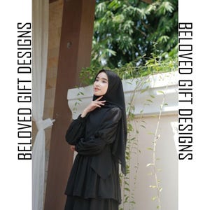 Op de afbeelding: Een vrouw draagt een zwarte jurk en hijab. De jurk heeft lange mouwen en een gelaagde rok. De tekst "BELOVED GIFT DESIGNS" is verticaal aan beide zijden van de afbeelding afgedrukt.