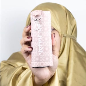 May include: A light pink rectangular case with a geometric pattern and Arabic text. The case has a silver-colored carabiner clip and a key ring. The background is a light gold color.