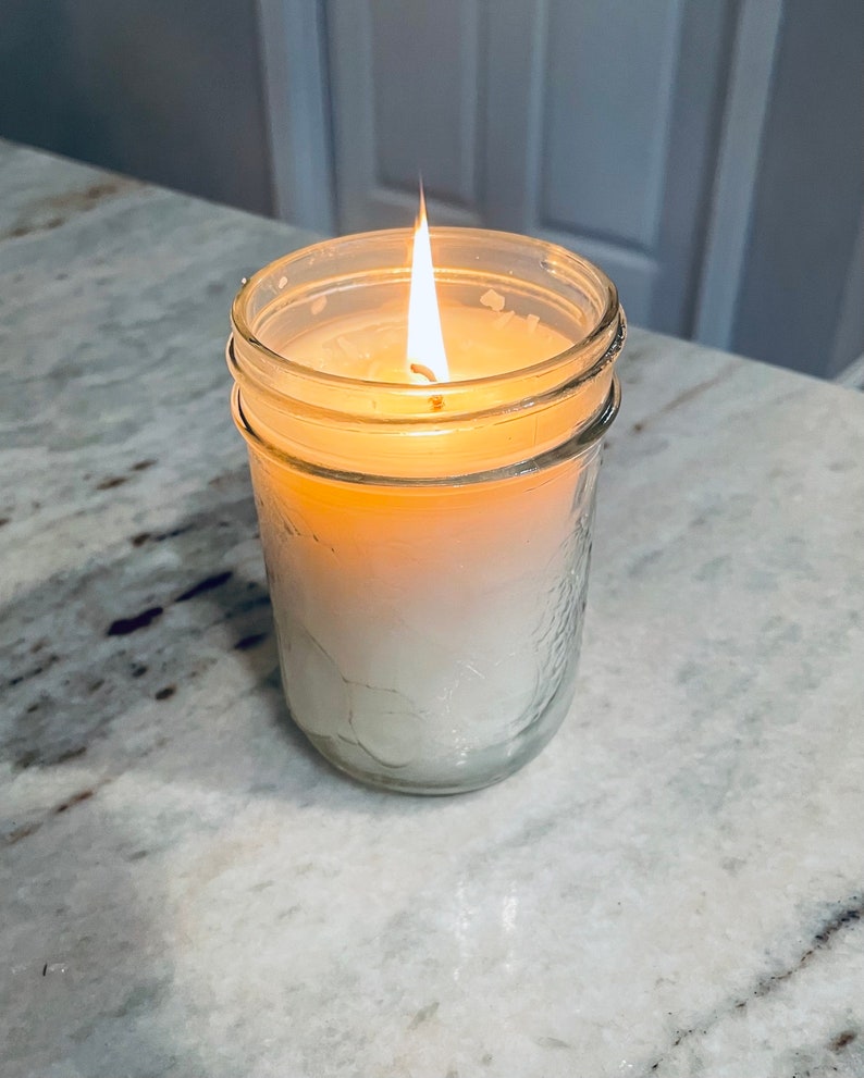 May include: A white candle burning in a clear glass jar with a textured surface. The candle is on a white marble countertop.