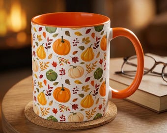 Può includere: Una tazza in ceramica bianca con interno e manico di un arancione vivace, caratterizzata da un motivo ripetuto di zucche arancioni, verdi e color crema, intervallate da piccole bacche rosse e foglie autunnali nei toni del giallo, arancione e marrone.