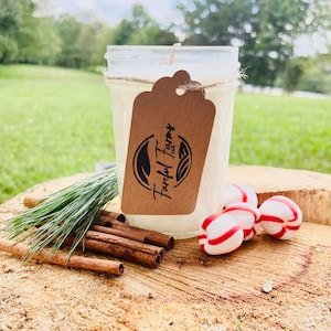 A white candle in a clear mason jar with a brown tag sits on a tree stump, with pine needles, cinnamon sticks, and three red and white candies.