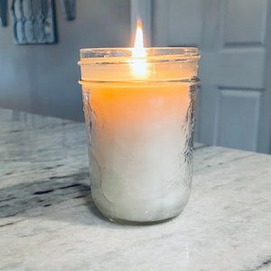A lit white candle in a clear mason jar with a textured surface sits on a speckled white countertop, with a blurred background.