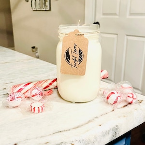 A white candle in a clear mason jar with a brown tag sits on a speckled white countertop, surrounded by red and white candy canes and wrapped candies.