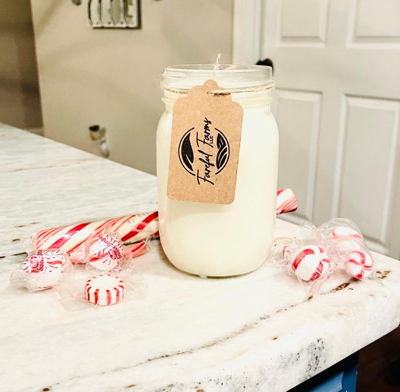 A white candle in a clear mason jar with a brown tag sits on a speckled white countertop, surrounded by red and white candy canes and wrapped candies.