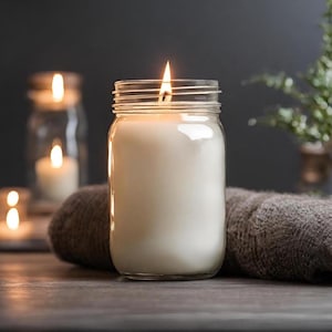 May include: A white candle in a clear glass jar with a single wick burning. The candle is sitting on a wooden surface with a brown towel beside it.