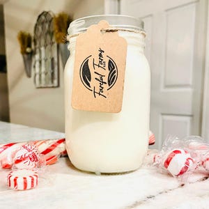 A white candle in a clear mason jar with a brown tag sits on a speckled white countertop, with red and white candies scattered around its base.