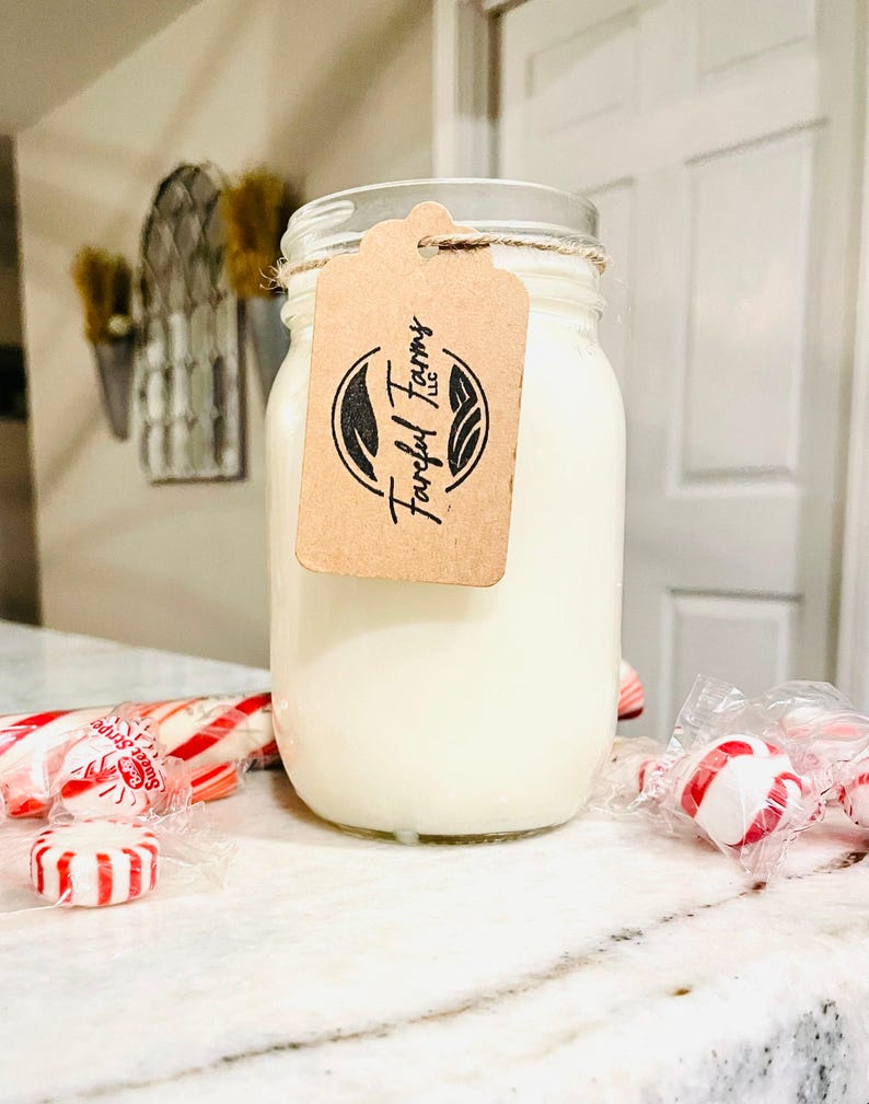 A white candle in a clear mason jar with a brown tag sits on a speckled white countertop, with red and white candies scattered around its base.