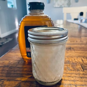 May include: A clear glass jar with a silver lid filled with white crystals sits on a wooden table next to a glass bottle of honey with a black lid.