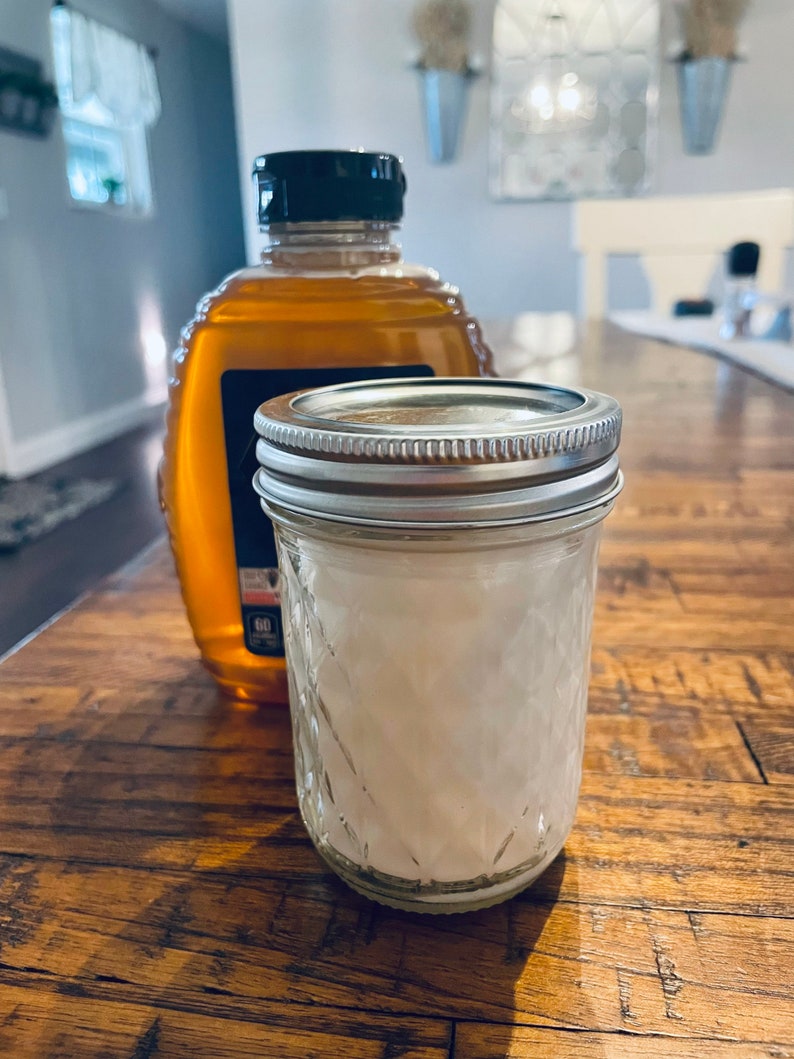 May include: A clear glass jar with a silver lid filled with white crystals sits on a wooden table next to a glass bottle of honey with a black lid.