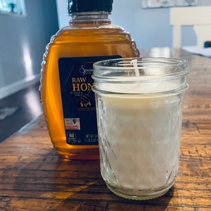 May include: A clear glass jar candle with a white wax interior and a white wick. The candle is sitting on a wooden table next to a bottle of honey labeled "Special Select Raw Honey".