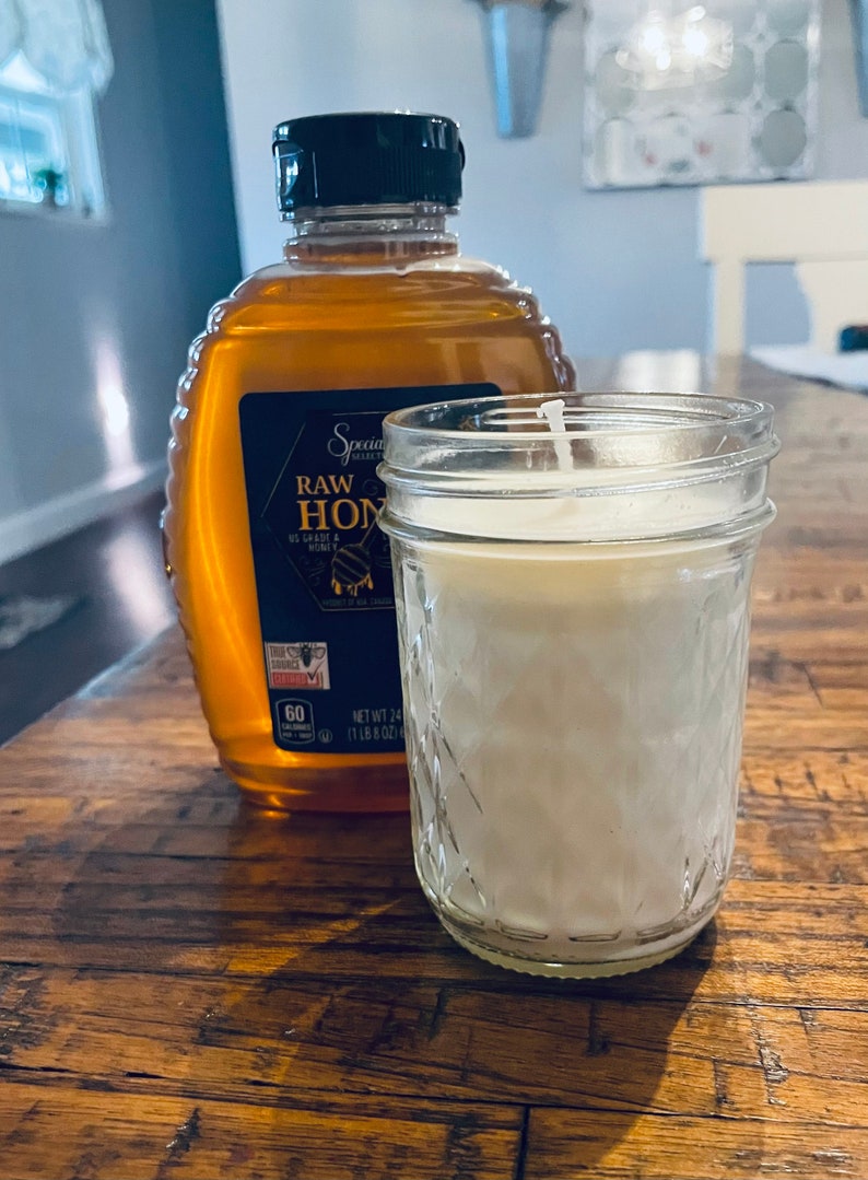 May include: A clear glass jar candle with a white wax interior and a white wick. The candle is sitting on a wooden table next to a bottle of honey labeled "Special Select Raw Honey".