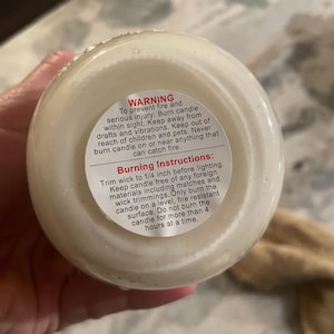 The bottom of a white candle in a jar, held by a hand, shows a white warning label with black text about candle safety and burning instructions.