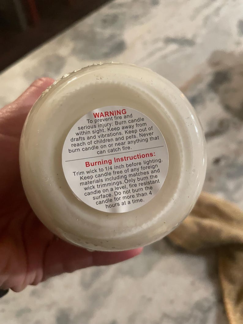 The bottom of a white candle in a jar, held by a hand, shows a white warning label with black text about candle safety and burning instructions.