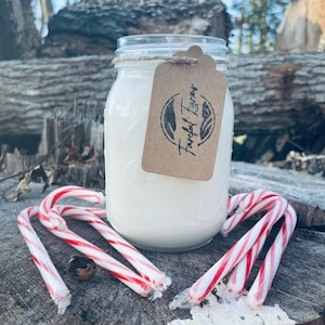 A white candle in a clear mason jar with a brown tag sits on a tree stump, surrounded by three red and white candy canes.