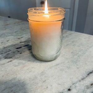 May include: A clear glass jar candle with a white wax and a lit wick. The candle is sitting on a white countertop.