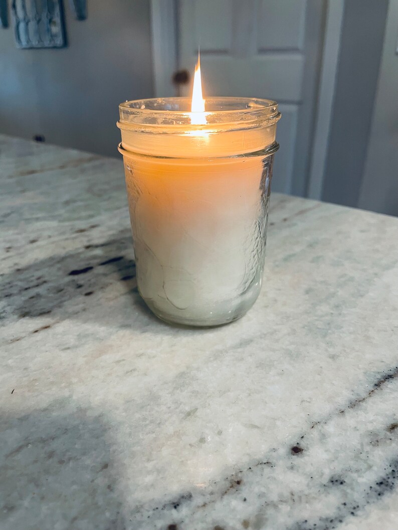 May include: A clear glass jar candle with a white wax and a lit wick. The candle is sitting on a white countertop.