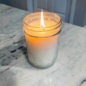 A lit white candle in a clear mason jar with a textured surface sits on a speckled white countertop, with a blurred white cabinet in the background.