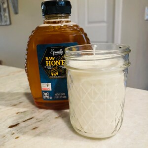 May include: A clear glass jar candle with a white wax interior and a diamond pattern on the outside. The candle is sitting on a white countertop next to a bottle of raw honey.