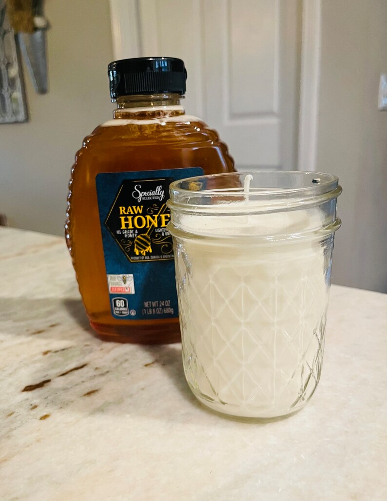 May include: A clear glass jar candle with a white wax interior and a diamond pattern on the outside. The candle is sitting on a white countertop next to a bottle of raw honey.