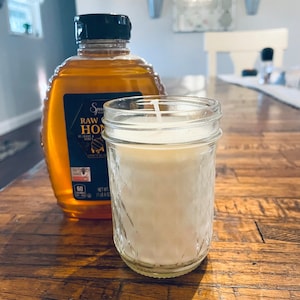 May include: A clear glass jar candle with a white wax interior and a black lid. The candle is sitting on a wooden table next to a bottle of raw honey.