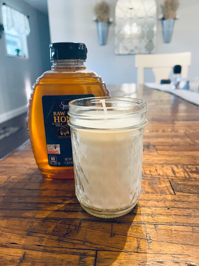 May include: A clear glass jar candle with a white wax interior and a black lid. The candle is sitting on a wooden table next to a bottle of raw honey.